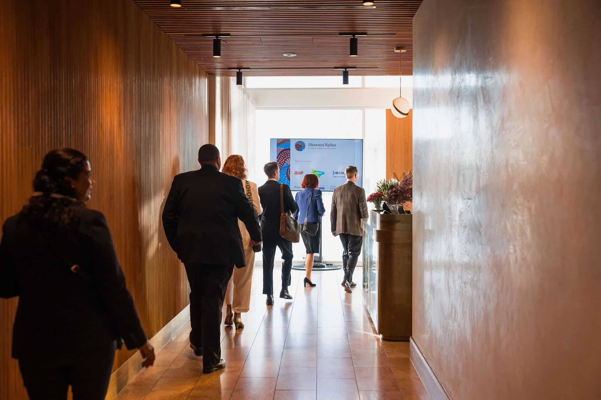 Professionals walk down a wood-paneled hallway toward a presentation screen in a bright lobby.