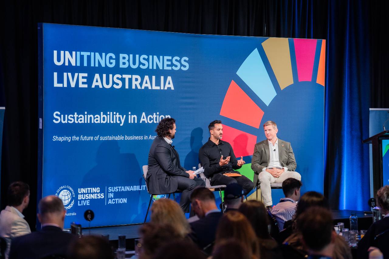 Panel discussion on stage with a blue backdrop reading 'Uniting Business Live Australia' and 'Sustainability in Action'; three men seated in conversation, audience in foreground.